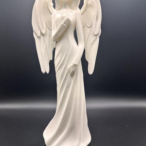 May include: A white angel figurine with outstretched wings. The angel is wearing a long dress and holding a heart. The figurine is standing on a dark surface.
