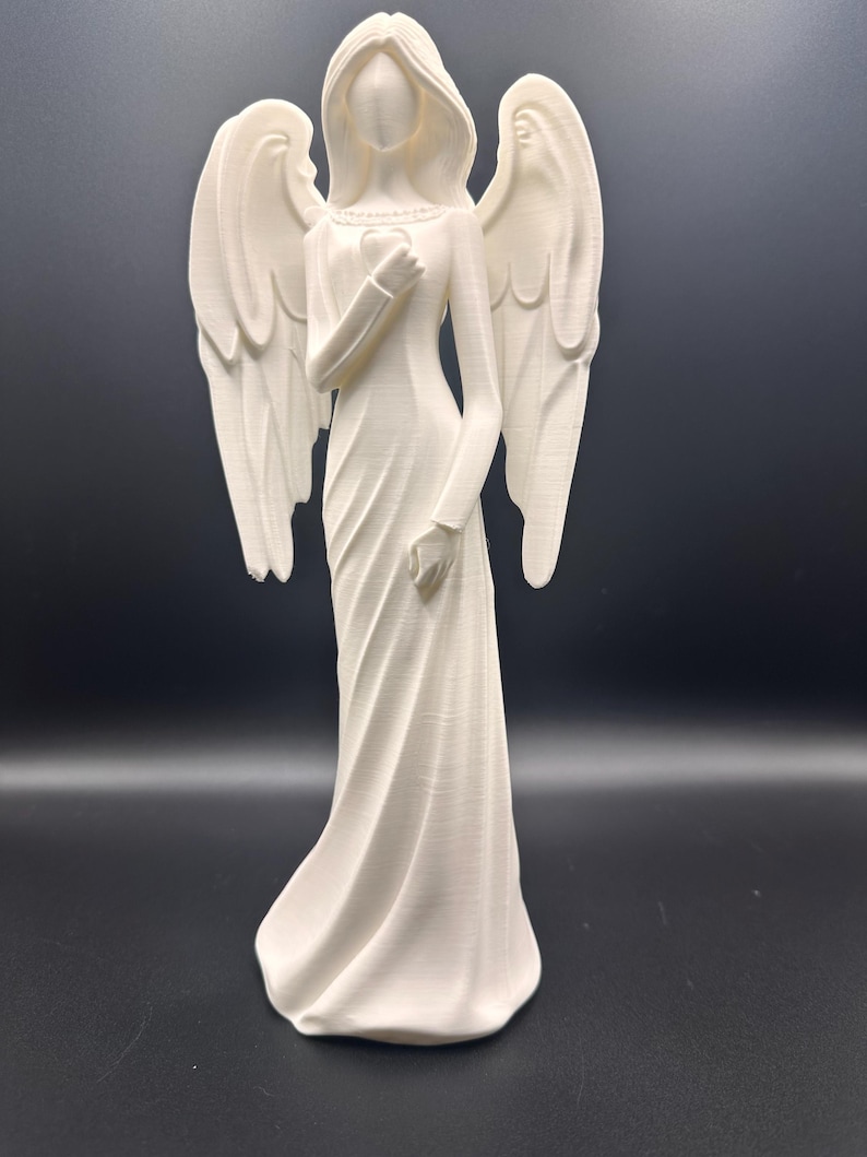 May include: A white angel figurine with outstretched wings. The angel is wearing a long dress and holding a heart. The figurine is standing on a dark surface.
