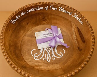 May include: A wooden bowl with a beaded rim and the inscription "Under the protection of Our Blessed Mother." A card tied with a purple ribbon rests inside the bowl. The bowl has a decorative white design.