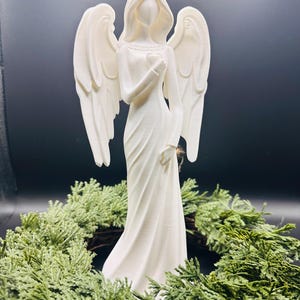 May include: A white angel figurine with outstretched wings, holding a heart, stands atop a green wreath. The angel is dressed in a flowing gown. The background is a dark, solid color.