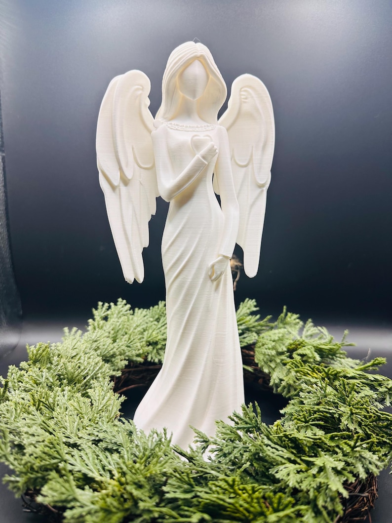 May include: A white angel figurine with outstretched wings, holding a heart, stands atop a green wreath. The angel is dressed in a flowing gown. The background is a dark, solid color.