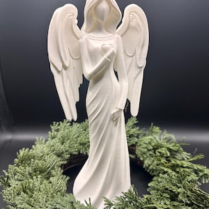 May include: A white angel figurine with outstretched wings, holding a heart. The angel is wearing a long dress and is surrounded by a green wreath. The figurine is made of a smooth material and has a serene expression.
