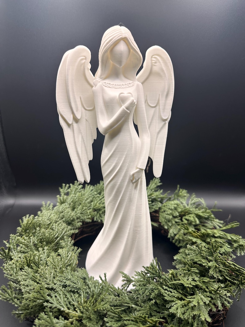 May include: A white angel figurine with outstretched wings, holding a heart. The angel is wearing a long dress and is surrounded by a green wreath. The figurine is made of a smooth material and has a serene expression.