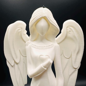 May include: A white angel figurine with flowing hair and large wings. The angel is holding a heart in its hand. The figurine has a detailed design and is set against a black background. The angel's dress has a detailed neckline.