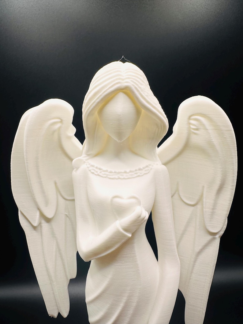 May include: A white angel figurine with flowing hair and large wings. The angel is holding a heart in its hand. The figurine has a detailed design and is set against a black background. The angel's dress has a detailed neckline.