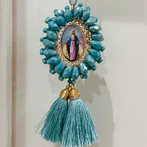 Op de afbeelding: Een religieuze hanger met een ovale afbeelding van een heilige, omringd door turquoise kralen. De hanger heeft een goudkleurig frame en is versierd met twee turquoise kwastjes. Hij hangt aan een bijpassend turquoise koord.