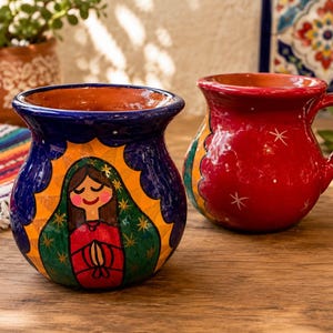 May include: Two round ceramic mugs with a glossy finish. The front mug is dark blue with a hand-painted figure in a green and red robe surrounded by a yellow sunburst. The back mug is solid red with small white star accents and a side handle.