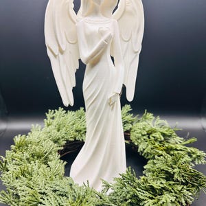May include: A white angel figurine with outstretched wings, holding a heart, stands in front of a green wreath. The angel is wearing a long, flowing gown. The background is a dark, solid color.