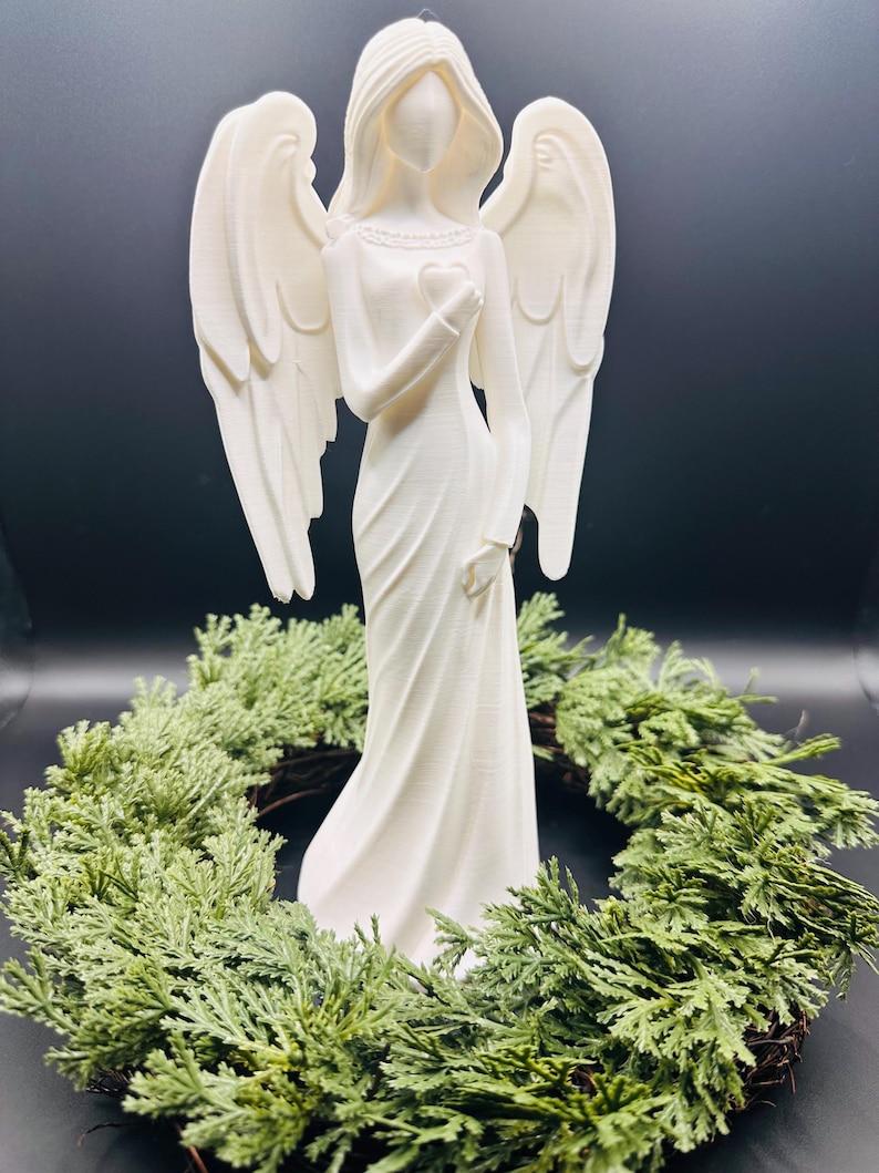 May include: A white angel figurine with outstretched wings, holding a heart, stands in front of a green wreath. The angel is wearing a long, flowing gown. The background is a dark, solid color.