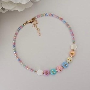 May include: A beaded necklace featuring a strand of small, multi-colored pastel faceted beads. The necklace includes white star-shaped beads and round, pastel-colored letter beads that spell out the name OLIVIA, finished with a gold-tone metal clasp.