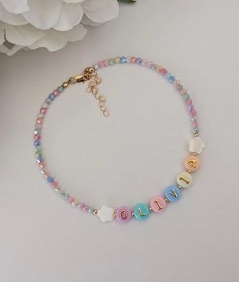 May include: A beaded necklace featuring a strand of small, multi-colored pastel faceted beads. The necklace includes white star-shaped beads and round, pastel-colored letter beads that spell out the name OLIVIA, finished with a gold-tone metal clasp.