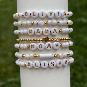 Peut inclure: Un empilement de sept bracelets de perles avec des perles rondes blanches, des perles intercalaires métalliques dorées et des perles lettres blanches épelant OLIVIA, MAMA, NOAH et ALISHA. Un bracelet inclut un petit cœur doré et un tube blanc.