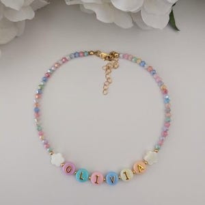 May include: A beaded necklace featuring small, multi-colored faceted beads and round, pastel-colored letter beads spelling the name OLIVIA. The necklace includes two white flower-shaped beads, gold-toned metal accents, and a gold-toned lobster clasp.