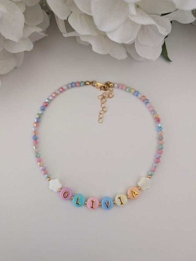 May include: A beaded necklace featuring small, multi-colored faceted beads and round, pastel-colored letter beads spelling the name OLIVIA. The necklace includes two white flower-shaped beads, gold-toned metal accents, and a gold-toned lobster clasp.