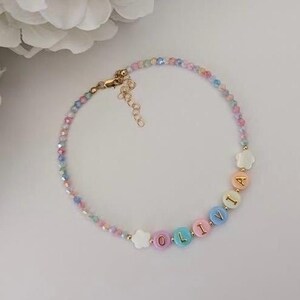 May include: A beaded bracelet featuring small, multi-colored faceted beads and larger, round, pastel-colored letter beads spelling the name OLIVIA. The bracelet includes two white flower-shaped beads and a gold-toned metal lobster clasp with an extender chain.