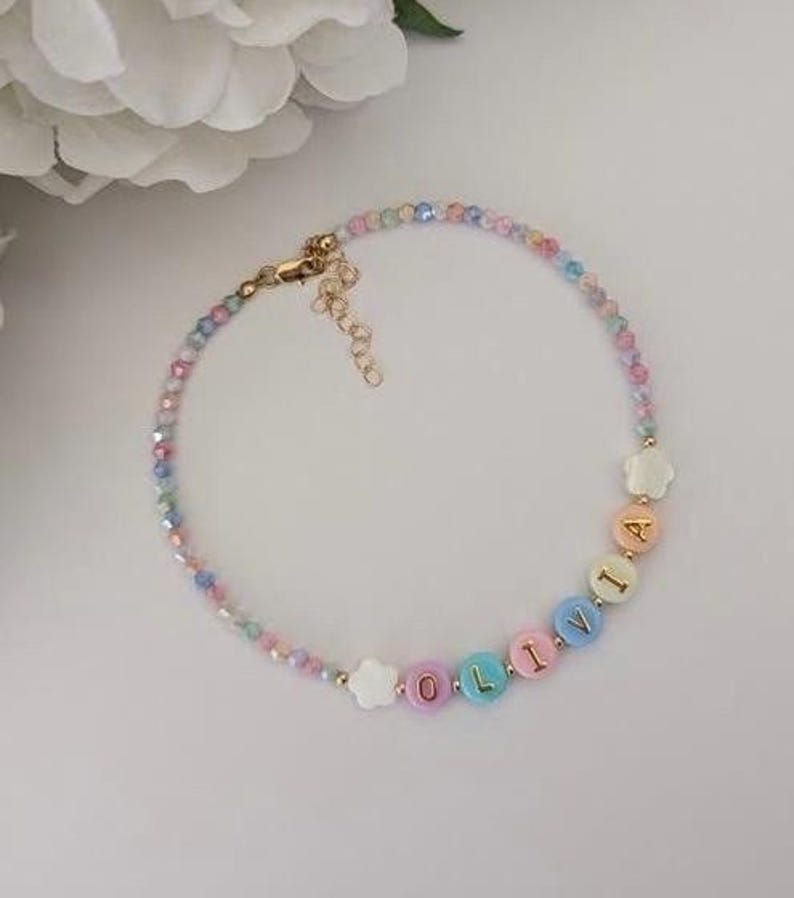 May include: A beaded bracelet featuring small, multi-colored faceted beads and larger, round, pastel-colored letter beads spelling the name OLIVIA. The bracelet includes two white flower-shaped beads and a gold-toned metal lobster clasp with an extender chain.