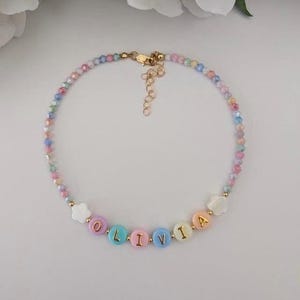 May include: A beaded necklace featuring a strand of small, multi-colored faceted beads in pastel shades. The center includes round, flat beads spelling the name OLIVIA in gold letters, flanked by two white flower-shaped beads and a gold-tone metal clasp.