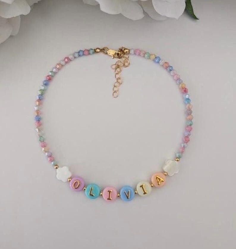 May include: A beaded necklace featuring a strand of small, multi-colored faceted beads in pastel shades. The center includes round, flat beads spelling the name OLIVIA in gold letters, flanked by two white flower-shaped beads and a gold-tone metal clasp.