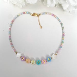May include: A beaded necklace featuring small, multi-colored pastel beads and a central section with round, colorful beads spelling the name OLIVIA in gold letters. The design includes two white flower-shaped beads and a gold-tone metal clasp.