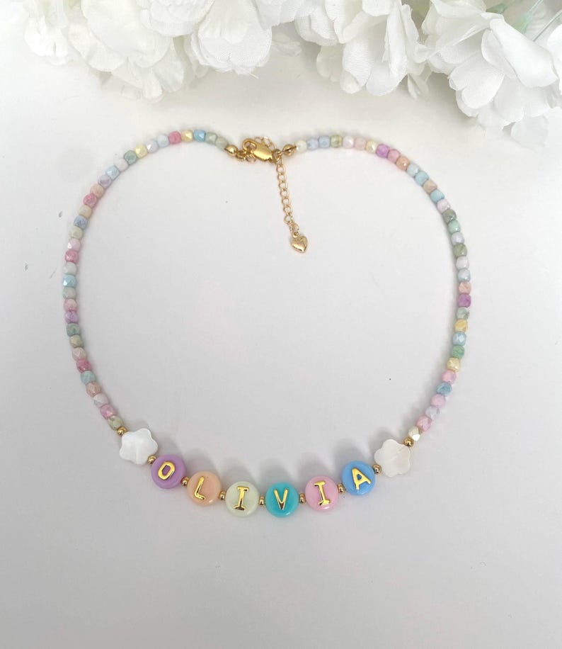 May include: A beaded necklace featuring small, multi-colored pastel beads and a central section with round, colorful beads spelling the name OLIVIA in gold letters. The design includes two white flower-shaped beads and a gold-tone metal clasp.