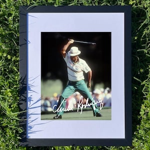 以下が含まれることがあります： ゴルフのスイング中のゴルファーの額装写真。ゴルファーは白い帽子、白と緑のストライプのシャツ、緑のズボンを着用しています。画像は緑色の背景で、ゴルファーのサインが見えます。