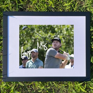 May include: Framed photograph of a golfer mid-swing, set against a green background. The image is in a black frame, resting on a bed of green grass. The golfer is wearing a cap and a gray shirt. The photograph has a signature in white.