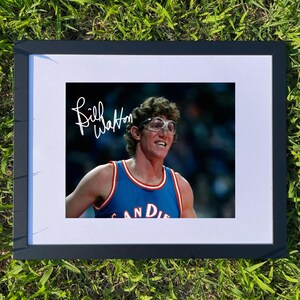 May include: Framed photograph featuring a smiling man in a blue and red basketball jersey. The jersey reads "San Diego". The photo is signed "Bill Walton" in white script. The frame is black.