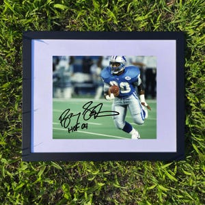 May include: Framed photograph of a football player in a blue uniform, running with the ball. The image is signed and includes the text "HOF 04". The frame is black, and the background is green grass.
