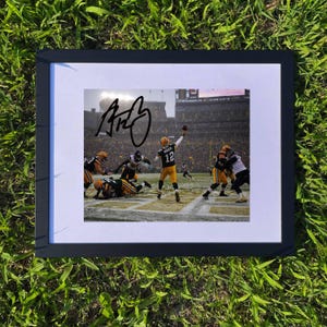 May include: Framed photograph of a football game featuring a player in a yellow and green uniform throwing a ball. The photo is signed with a black marker. The frame is black and the photo is on a white mount.