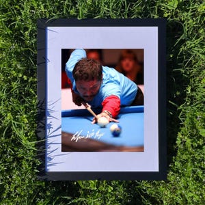 May include: Framed photograph of a man playing billiards, framed in black. The man is wearing a blue shirt with red sleeves, focused on aiming with a cue stick. The pool table is blue, with a white and a striped ball visible.