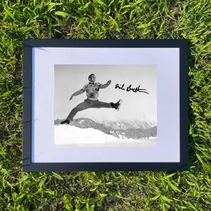 Może przedstawiać: Oprawione czarno-białe zdjęcie mężczyzny jeżdżącego na łyżwach, w trakcie skoku, z ośnieżonym górskim tłem. Zdjęcie jest podpisane "Dick Button" w prawym górnym rogu. Rama jest czarna, a zdjęcie jest umieszczone na białym passe-partout.