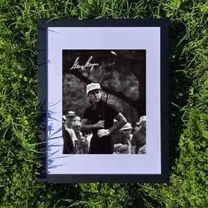 May include: Framed black and white photograph of a golfer in action, holding a golf club. The image is signed by the golfer, with a crowd of people in the background. The frame is black, and the photograph is set against a white mat.