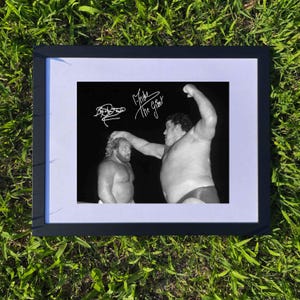 Könnte beinhalten: Gerahmtes Schwarz-Weiß-Foto mit zwei Wrestlern, von denen einer den anderen überragt. Das Bild ist mit den Namen "Andre The Giant" und einer weiteren Unterschrift versehen. Der Rahmen ist schwarz, mit einem weißen Passepartout, auf grünem Gras.