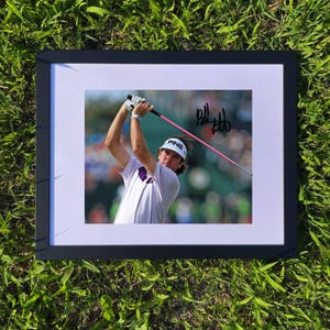 May include: Framed photograph of a golfer in action, swinging a pink golf club. The golfer is wearing a white shirt and a white visor with the word "PING" on it. The photograph is signed with the name "Bill Hubb". The frame is black.