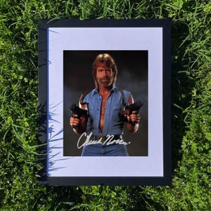 Puede incluir: Impresión enmarcada con Chuck Norris sosteniendo dos pistolas. La imagen muestra a Norris con camisa vaquera y vaqueros, con su firma en la parte inferior. La impresión está enmarcada en un marco negro con un passe-partout blanco.