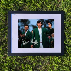 Op de afbeelding: Een ingelijste foto met twee mannen in groene blazers, waarvan één zwaait. De foto is geplaatst op een witte passe-partout en in een zwart frame. De afbeelding ligt op groen gras.