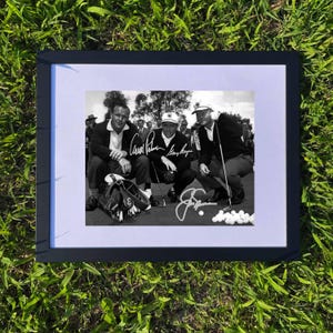 May include: Black and white framed photograph featuring three golfers, each with a signature. The golfers are seated or crouching on a golf course, with golf balls and a golf bag visible. The frame is black, and the background is green grass.