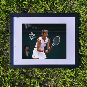 Puede incluir: Fotografía enmarcada de una tenista con un vestido de tenis blanco con detalles en rojo y naranja, sosteniendo una raqueta de tenis. La fotografía está firmada en la esquina superior izquierda. El marco es negro y el fondo es hierba verde.