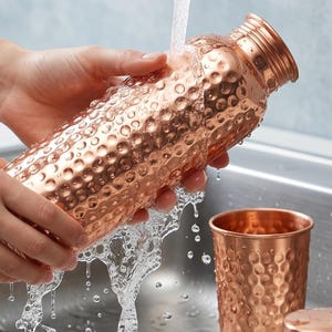 May include: A copper water bottle with a textured, dimpled surface is being rinsed under a faucet. The bottle is cylindrical with a screw-on cap. A matching copper cup and lid are also visible. The text "CLEAN & REFRESH" is in the background.