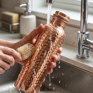 May include: A copper water bottle with a textured surface is being cleaned with a brush under running water. The bottle has a screw-on cap and a hammered design. The bottle is in a kitchen sink.