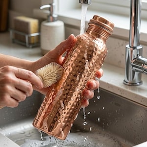 Puede incluir: Una botella de agua de cobre con una superficie texturizada se limpia con un cepillo bajo agua corriente. La botella tiene una tapa de rosca y un diseño martillado. La botella está en un fregadero de cocina.