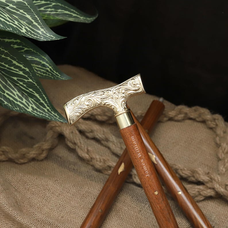 K&ouml;nnte beinhalten: Dekorative Gehstock mit einem goldfarbenen, verzierten Griff und einem dunkelbraunen Holzschaft. Der Griff weist aufwendige Blumenmuster auf. Der Schaft ist mit dem Namen "James William" graviert. Ein klassisches Accessoire.