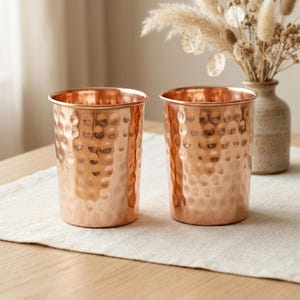 May include: Two copper-colored drinking glasses with a hammered texture. The glasses have a cylindrical shape and a smooth rim. They are sitting on a beige linen runner on a wooden table. A vase with dried flowers is in the background.