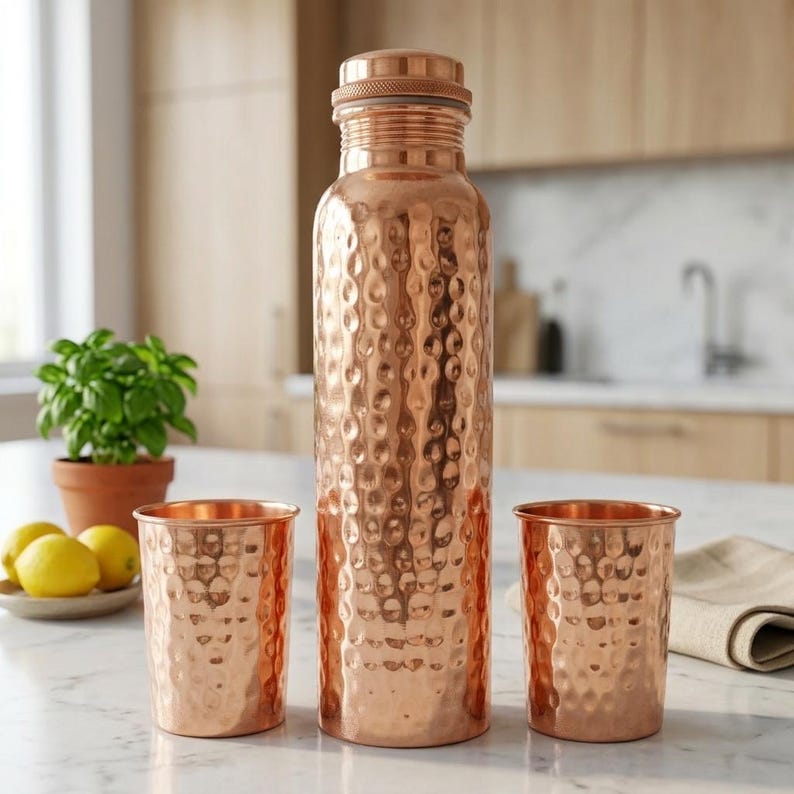 May include: A copper water bottle with a textured, hammered finish stands next to two matching copper cups. The bottle has a screw-on lid. The cups and bottle are on a marble surface, with a small plate of lemons and a potted plant in the background.