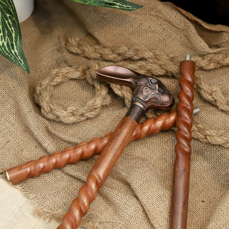 May include: A wooden walking cane with a bronze rabbit head handle. The cane has a twisted design and a bronze ferrule. The cane is resting on a burlap surface with rope.