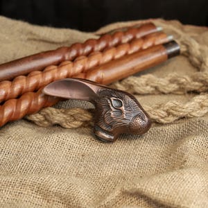 May include: A decorative walking cane with a bronze-colored rabbit head handle. The cane shaft is made of dark brown wood with a spiral design. The background features a burlap texture and rope.