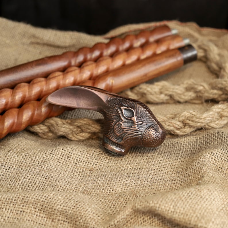 May include: A decorative walking cane with a bronze-colored rabbit head handle. The cane shaft is made of dark brown wood with a spiral design. The background features a burlap texture and rope.