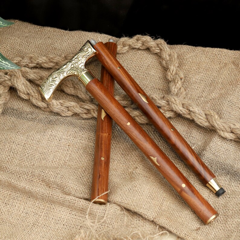 K&ouml;nnte beinhalten: Ein h&ouml;lzerner Spazierstock mit einem dekorativen Messinggriff und Akzenten. Der Griff weist ein verziertes Blumendesign auf. Der Stock ist braun mit Messingdetails und einer schwarzen Gummispitze. Der Stock ist etwa 91 cm lang.