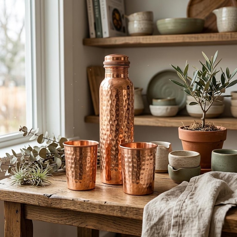May include: A copper water bottle and two matching copper tumblers sit on a wooden table. The bottle has a textured surface and a screw-on cap. The tumblers have a similar hammered texture. A small potted plant and other kitchenware are in the background.