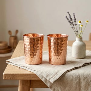 May include: Two copper-colored drinking glasses with a hammered texture sit on a wooden table. A small white vase with lavender and daisies is to the right. The glasses are cylindrical and have a shiny finish.